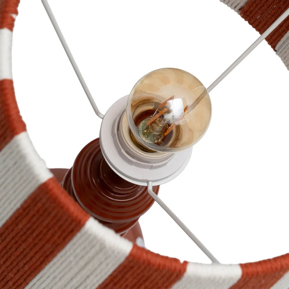 Close-up of a paper rope lampshade with alternating rust red and white stripes, showing a clear glass light bulb mounted in a white socket on a dark brown metal base | KULORI