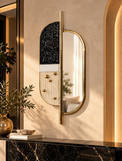 An asymmetrical modern wall mirror with a gold frame, featuring a black glittery arch on one side and a vertical gold rod with small gold knobs on the other, mounted on a textured beige wall in a warm, sunlit interior  | KULORI Malta & Gozo