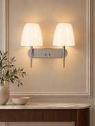 A modern two-light wall sconce with white tapered shades and a brushed silver metal base, mounted on a beige wall above a marble console table with a vase of greenery KULORI Malta