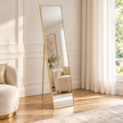 A tall floor mirror with a slim gold metal frame stands in a bright, elegant room with cream walls, wood floors, sheer curtains, and a matching cream armchair visible in the mirror reflection | KULORI Malta & Gozo