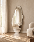 An organic-shaped floor mirror with a thin black frame stands against a textured beige wall in a bright, modern room. The mirror reflects a sunlit area with sheer curtains and a sofa, while the room features light wood flooring, a woven rug, and a textured cream boucle armchair An organic-shaped wall mirror with a soft, irregular outline | KULORI Malta & Gozo