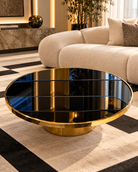 A modern round coffee table with a polished gold metal base and a black mirrored top, set in a luxury living room with a beige sofa and patterned rug | KULORI Malta & Gozo