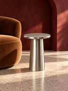 A modern cylindrical side table with a brushed silver metal finish, positioned next to a brown velvet armchair in a room with a terracotta archway and terrazzo floor | KULORI Malta & Gozo