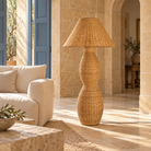 A tall floor lamp with a woven natural rattan shade and a sculptural double-bulb base in a warm beige tone, set in a sunlit stone interior next to a cream sofa  | KULORI Malta & Gozo