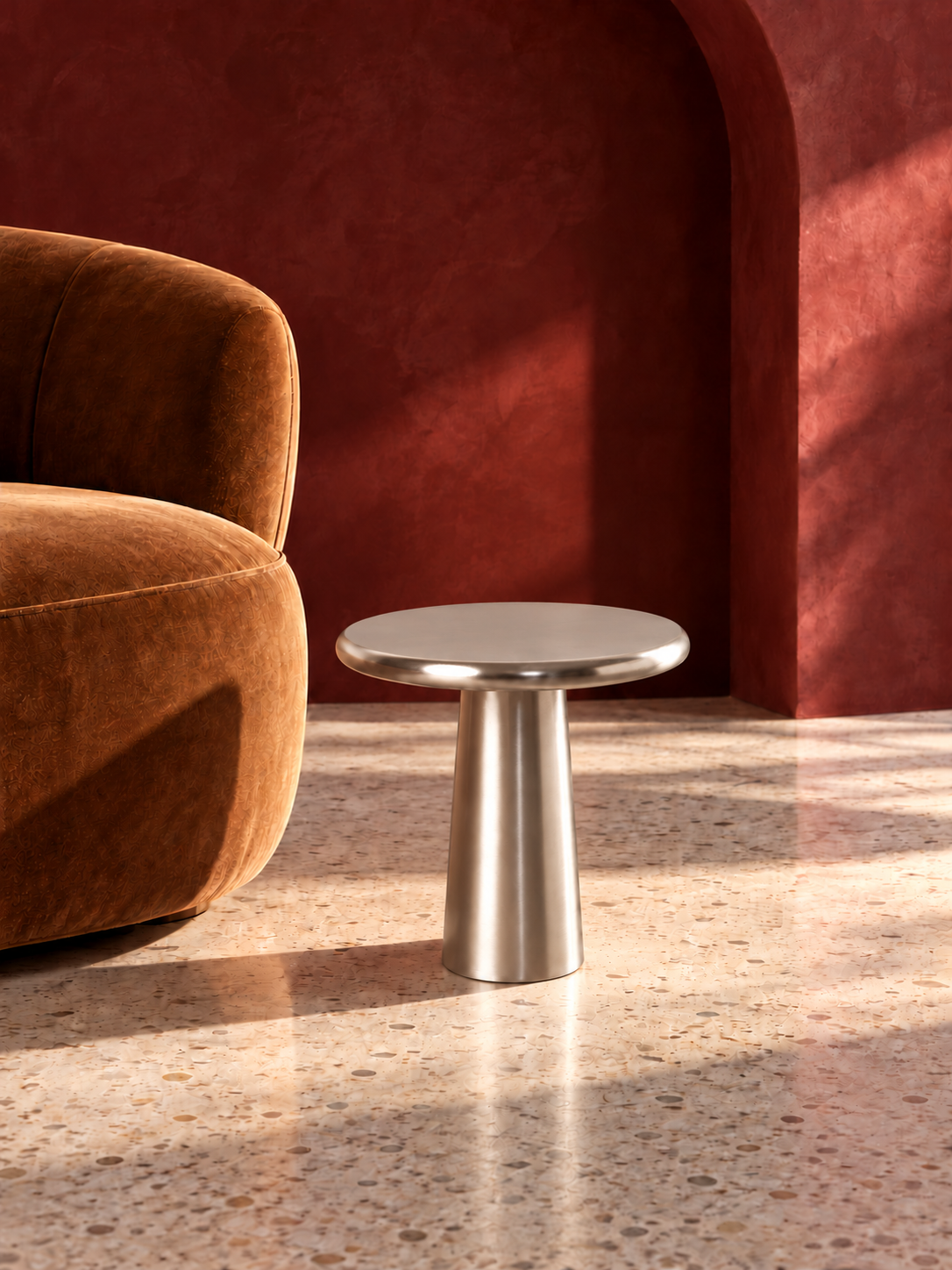 A small round side table with a brushed silver metal pedestal base, positioned next to a brown velvet armchair in a room with a textured dark red wall and terrazzo floor | KULORI Malta & Gozo
