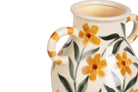 A close-up of a ceramic pitcher with a cream-colored glaze, decorated with yellow flowers and green leaves, featuring two loop handles
