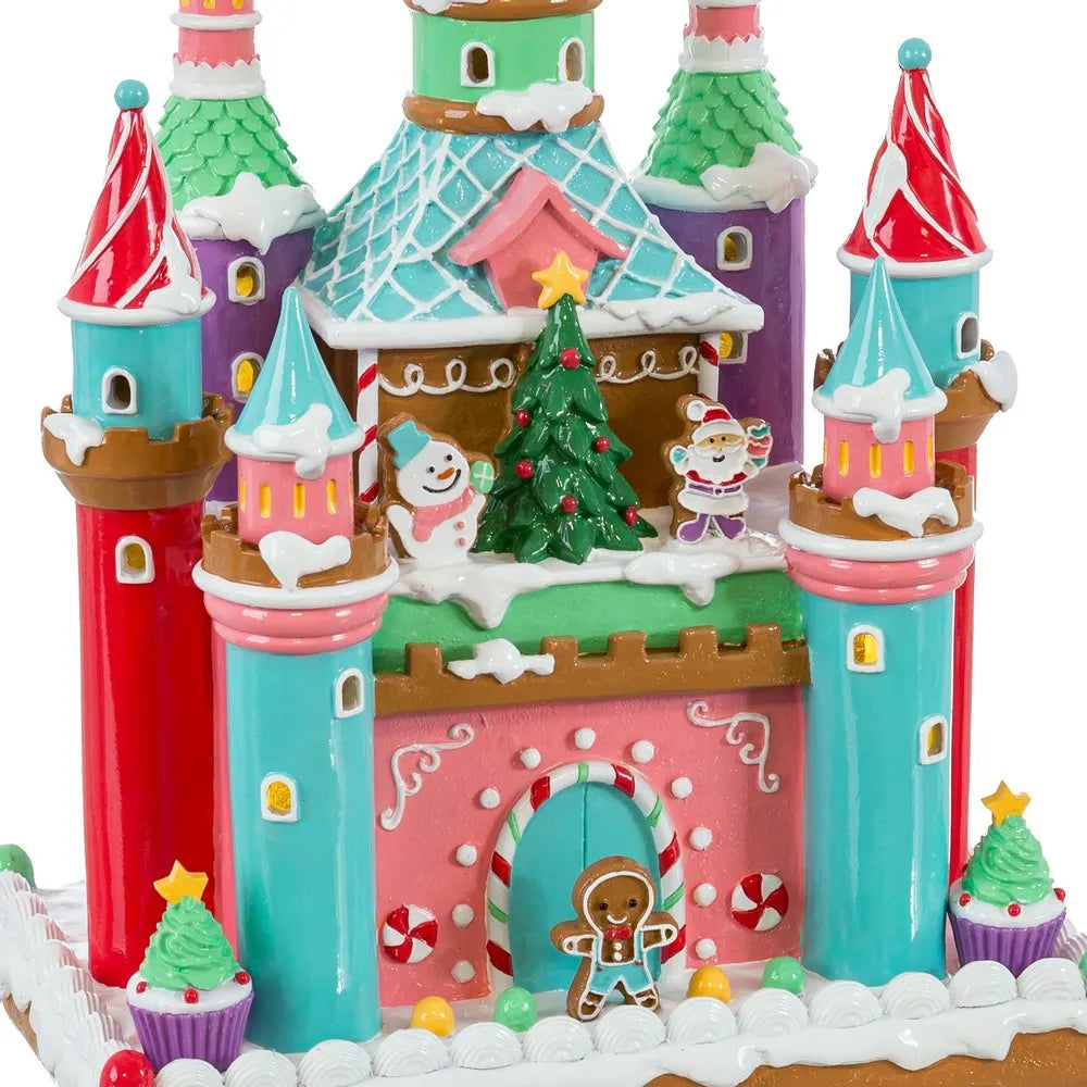 A colorful gingerbread castle decoration with snowy details, decorated with candy cane arches, a small green Christmas tree, a snowman, and a gingerbread person, shown on a white background.