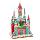 A colorful gingerbread castle decoration with red, pink, teal, green, and white icing-like details, snow-dusted edges, and small holiday figures and trees on a white background.