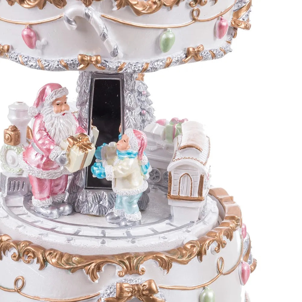 A close-up of a decorative Christmas music box featuring Santa Claus, a child in winter clothing, a small train, ornaments, and ornate gold-toned scrollwork on a white base.