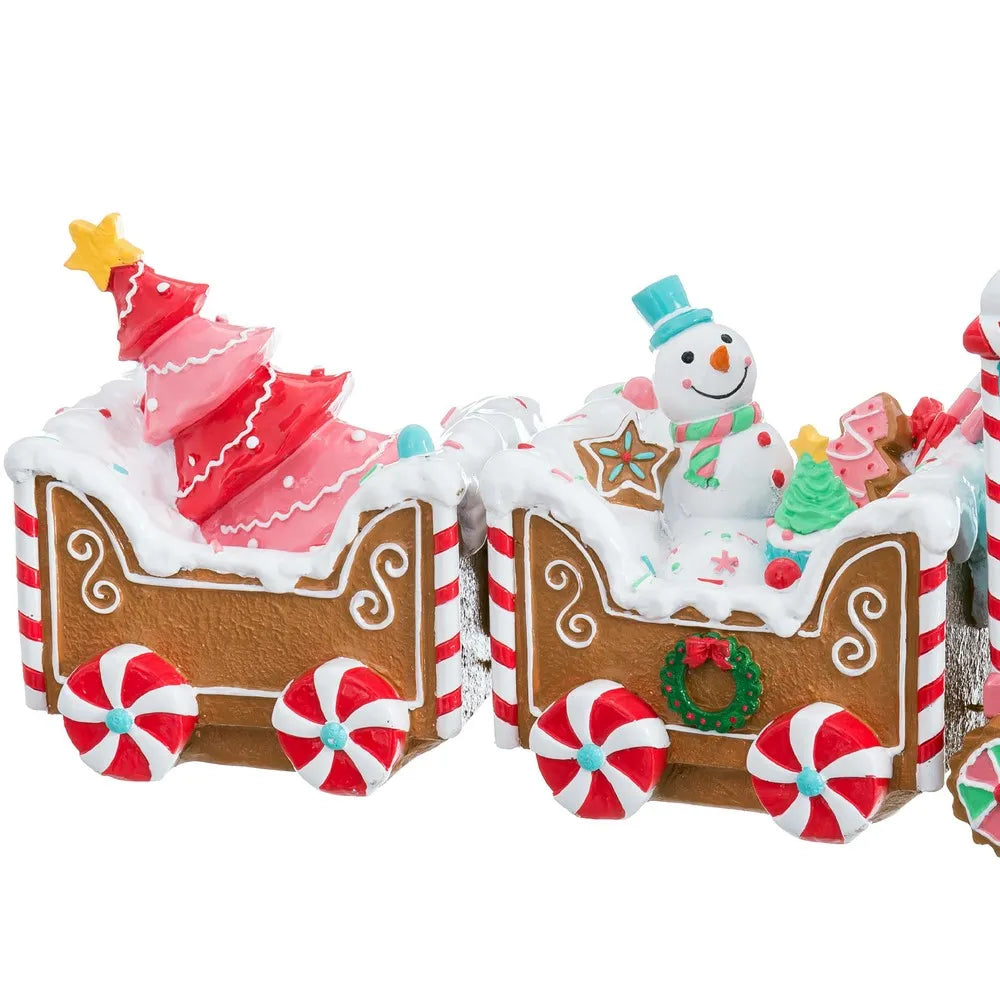 A festive Christmas gingerbread train decoration featuring two train cars decorated like gingerbread houses with white icing-like snow, candy cane wheels, a pink icing Christmas tree, a snowman figure, and small holiday treats, shown on a plain white background.
