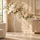 Two tall clear glass standees with wavy stems holding large floral arrangements in a sunlit living room. The arrangements feature white and blush pink flowers with green foliage, placed beside a sofa and console table.