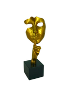 Gold mask sculpture with a hand covering its mouth on a black base against a white background | KULORI