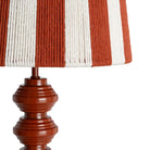 Close-up of a table lamp with a striped fabric shade in terracotta and off-white, mounted on a glossy reddish-brown turned metal base | KULORI